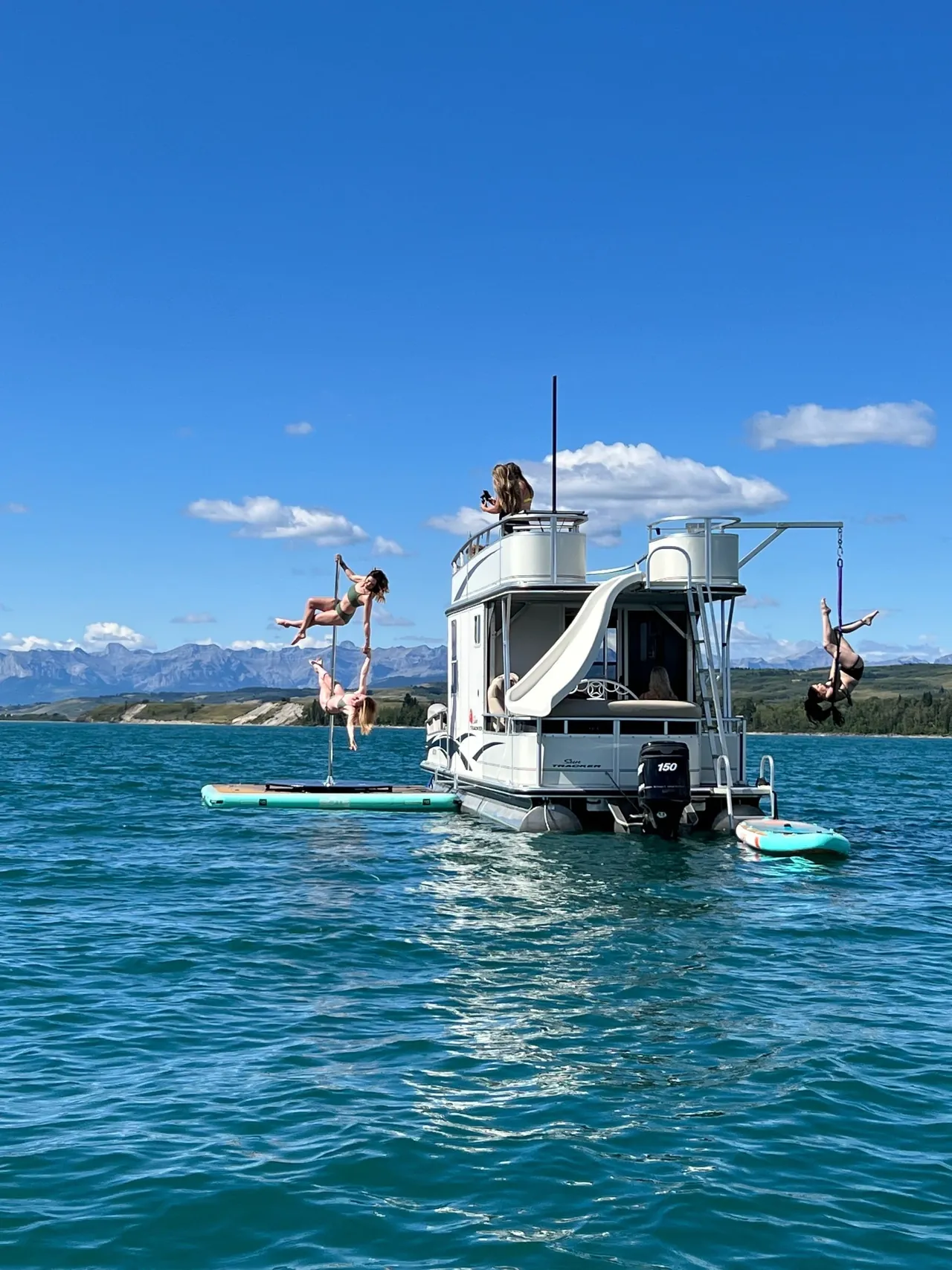 The R.Song AquaStudio double-decker pontoon boat on Ghost Lake with the water slide down, guests on the top deck and paddleboards alongside